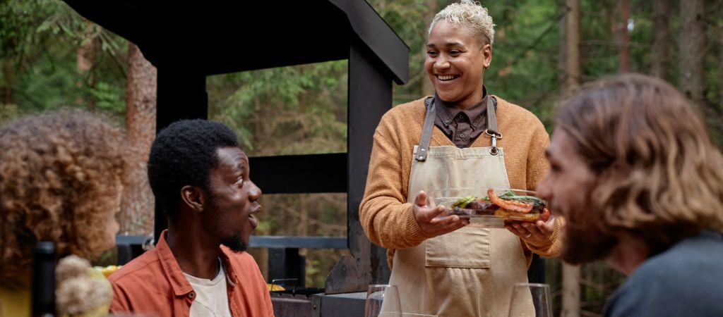 Guests at an outdoor table look with excitement as a smiling waiter brings out a fresh dish to serve. Friends Dining Outdoors