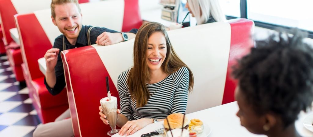 Friends laugh while seated in classic red and white booths at a 50s-style diner, with milkshakes and burgers on the tables. Friends Laughing at a Diner
