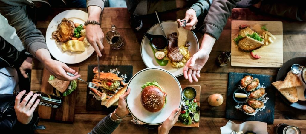 An overhead shot of a wooden table loaded with many dishes and hands passing plates for a large group meal. Group Dining Table at a Restaurant