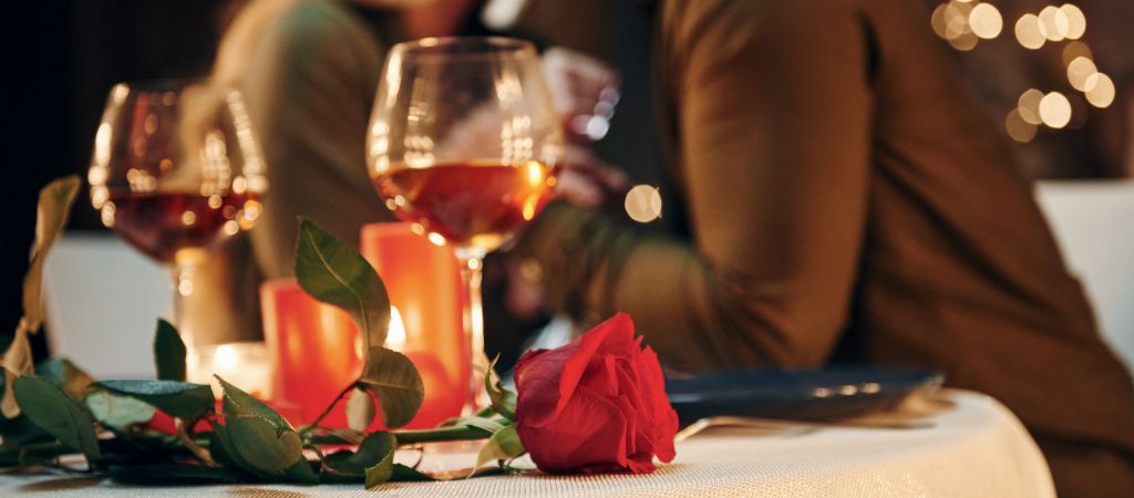 A rose and two glasses of wine sit on a cloth-dressed table, with a couple holding hands in the background. Wine Glasses at a Romantic Dinner