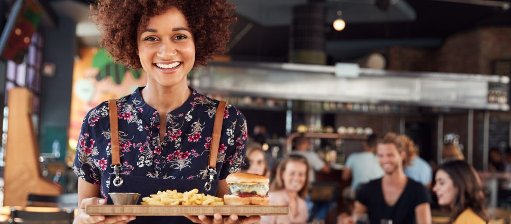 A waitress holds a serving board of classic American food while smiling at the camera, with a table of happy guests in the background. Portrait of a Waitress Serving Food