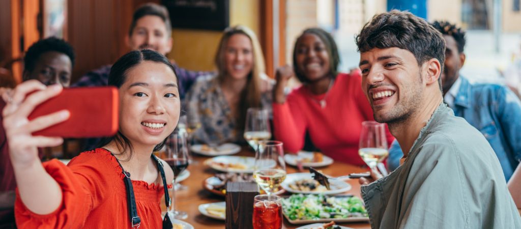 A young woman snaps a group selfie with smiling friends seated around a restaurant table loaded with dishes. Young Group Taking Selfie at a Restaurant