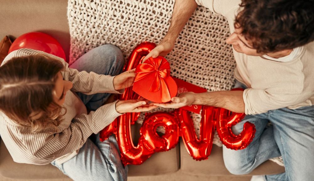 A top-down shot of a young couple sitting on a sofa with red balloons, one says "Love," and holding a heart-shaped red box with a bow.