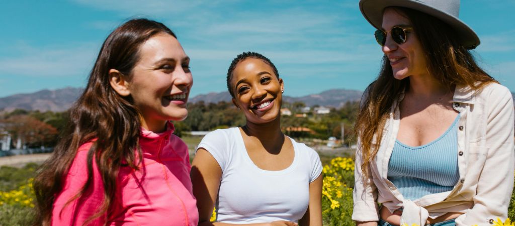 Three young women stand and smile in a field of yellow wildflowers on a sunny day, with mountains in the background and a blue sky overhead.