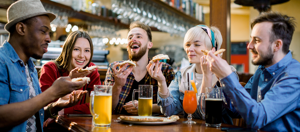 Young People Eating at a Restaurant A group of friends smile and eat pizza at a local brewery, with beer and cocktails on the table.
